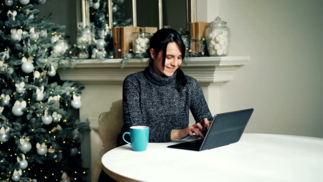 Chica-feliz-es-trabajar-con-la-laptop-en-día-sentado-en-la-mesa-cerca-de-árbol-de-año-nuevo-decorado-de-Navidad-y-escribir-mirando-a-la-pantalla-de-envío-de-correos-electrónicos-a-amigos.