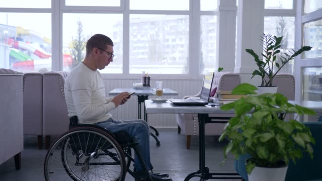 Freelancer-männliche-Behinderte-im-Rollstuhl-auf-Handy-und-Computer-Laptop-sitzen-an-einem-Tisch-in-einem-Café-eine-Brille