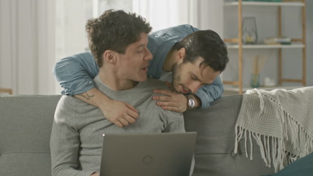 Linda-pareja-Gay-hombre-pasar-tiempo-en-casa.-Hombre-joven-trabaja-en-un-ordenador-portátil,-su-compañero-viene-desde-atrás-y-suavemente-le-abraza.-Se-ríen-y-tocar-manos.-Habitación-tiene-un-Interior-moderno.