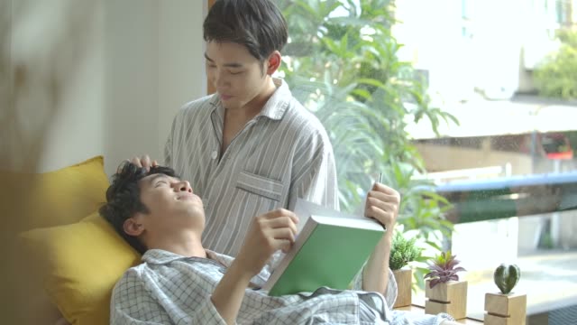 Asiático-gay-pareja-joven-sentada-en-el-alféizar-de-la-ventana-y-leyendo-el-libro-en-casa-en-la-mañana.