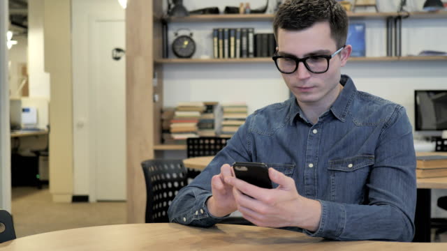 Joven-creativo-en-gafas-excitado-para-tener-éxito-al-Smartphone-utilizando