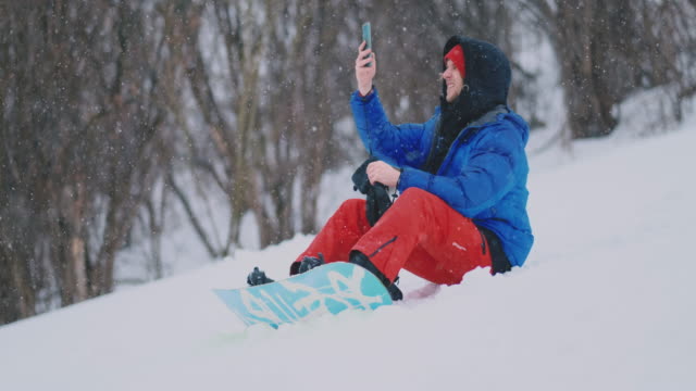 Männliche-Snowboarder-sitzen-auf-dem-Schnee-und-machen-Fotos-am-Telefon-schöne-Landschaft-des-Resorts-für-soziale-Netzwerke