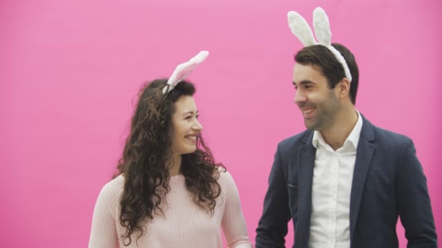Young-beautiful-handsome-happy-happy-couple-standing-on-a-pink-background.-At-the-same-time,-they-are-dressed-in-paschal-ears.-Sincere-smiles-make-the-movements-of-hares.-Easter-concept.