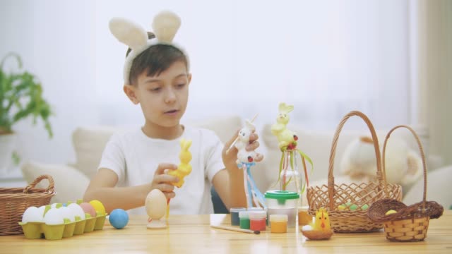Young-adorable-boy-is-sitting-at-the-table-full-of-Easter-decorations-and-is-playing-with-Easter-bunnies-in-his-hands.-Bunnies'-discussion.-Who-would-paint-an-egg-Bunny-chooses-green-colour.-Bunny-theatre.