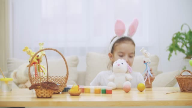Little-jouful-girl-with-bunny-ears-on-his-head-is-playing-on-the-wooden-table,-full-of-Easter-decorations.-Girl-is-playing-with-Easter-bunnies-and-an-yellow-egg,-isolated.