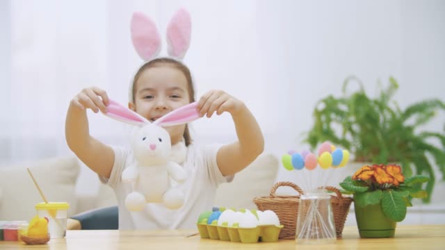 Little-cute-girl-is-having-fun-painting-with-Easter-bunny,-which-has-the-same-ears,-as-she-has.-Smiling-girl-is-expanding-bunny's-ears-and-then-is-hugging-it.-Girl-with-a-beauty-spot-at-her-face-and-is-smiling-widely,-sitting-at-the-wooden-table-with-East