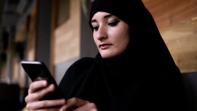 Cerca-de-la-mujer-musulmana-con-maquillaje-en-la-cafetería-usando-su-teléfono-inteligente,-chatear-en-línea-con-amigos-o-navegar-por-las-redes-sociales,-compartiendo-estilo-de-vida.-Disfrutando,-relajando-los-mensajes-de-lectura.-Mujer-vistiendo-hiyab-n