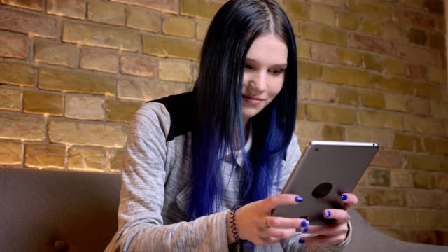 Closeup-shoot-of-young-pretty-caucasian-hipster-female-with-dyed-hair-texting-on-the-tablet-and-smiling-in-a-cozy-apartment-indoors