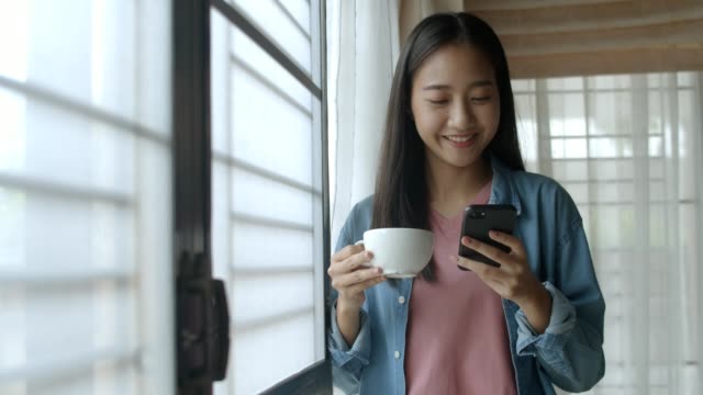 Attraktive-Porträt-lächelnde-junge-asiatische-Frau-trinken-Kaffee-und-tippen-Handy-und-scrollt-durch-Social-Media-Feed-in-Smartphone-neben-dem-Fenster-im-Home-Office-stehen.