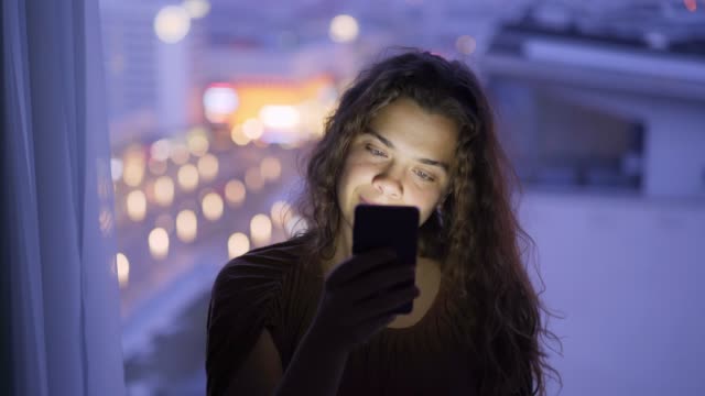 Mujer-de-primer-plano-usando-el-teléfono-contra-la-ventana-grande-en-el-crepúsculo