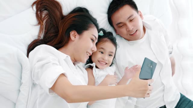 Asiatische-Familie-lacht-und-schaut-in-Smartphone,-während-auf-dem-Bett-im-Schlafzimmer-liegen