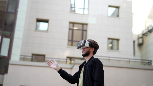 Panning-slow-motion-shot-of-young-man-in-vr-glasses-standing-outdoors-in-city-street-and-exploring-data-visualized-in-virtual-reality.-Man-swiping-and-magnifying-info
