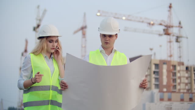 Two-builders-with-drawings-standing-on-the-background-of-buildings-under-construction-in-helmets-and-vests,-a-woman-talking-on-the-phone-with-the-customer