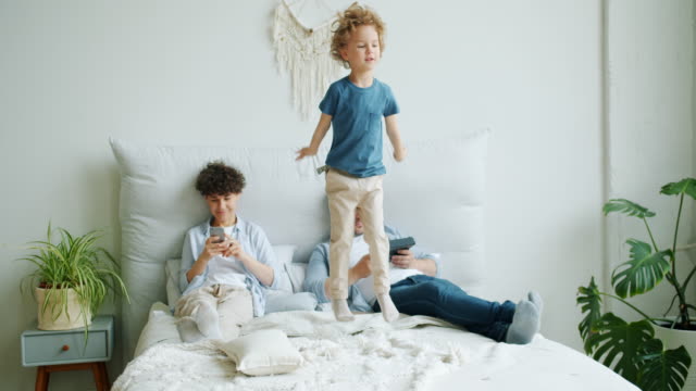 Fröhliches-Kind-springt-aufs-Bett,-während-Mutter-mit-Smartphone-Vater-halten-Tablet