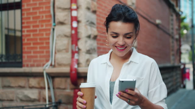 Slow-motion-of-beautiful-lady-using-smartphone-holding-coffee-walking-smiling