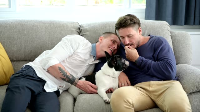 Gay-couple-relaxing-on-couch-with-dog.-Trying-to-mouth-feed-it.