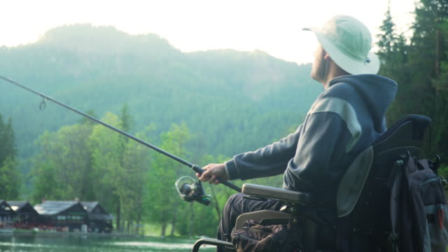 Resolución-4k-de-pescador-discapacitado-en-una-silla-de-ruedas-eléctrica-pescando-en-un-hermoso-lago-cerca-del-bosque-y-la-montaña-en-la-parte-posterior,-en-la-puesta-de-sol,-verano
