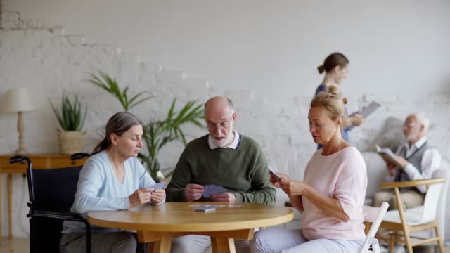 Tracking-Aufnahme-von-drei-Senioren,-die-Karten-spielen,-die-am-Tisch-im-Pflegeheim-sitzen.-Junge-Krankenschwester-im-Gespräch-mit-männlichen-alten-Patienten-Lesen-Buch-auf-Sofa-im-Hintergrund