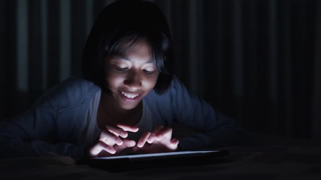 Asiática-niña-tocando-el-teclado-en-la-tableta-digital-moderna-en-la-cama-por-la-noche