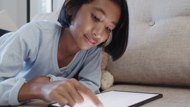 Asiat-des-kleinen-Mädchens-berührt-den-weißen-digitalen-Tablet-Bildschirm-und-lächeln,-während-sie-zu-Hause-auf-dem-Sofa-im-Wohnzimmer-liegen.