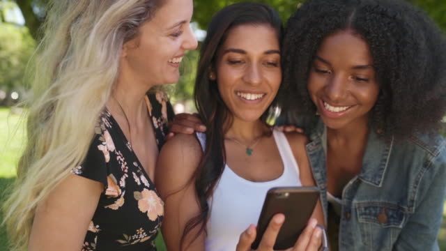 Fröhliche-drei-verschiedene-Freundinnen-hängen-im-Park-mit-Handy-und-teilen-digitale-Inhalte-auf-Social-Media-lächelnd-und-glücklich---Blick-auf-die-Handy-App