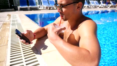 Hombre-guapo-usando-el-teléfono-en-la-piscina-del-hotel-y-refrescante-cóctel-frío.-Joven-con-gafas-de-sol-beber-bebida-haciendo-foto-para-la-red-social.-Hombre-relajante-en-el-complejo-durante-las-vacaciones-de-verano.-Cámara-lenta