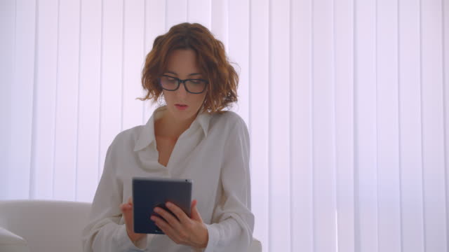 Closeup-portrait-of-adult-redhead-caucasian-businesswoman-in-glasses-using-the-tablet-looking-at-camera-smiling-happily-sitting-in-armchair-in-white-office