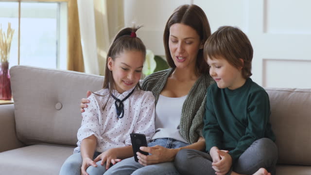 Glückliche-junge-Mama-unterrichtet-Kinder-mit-Handy-sitzen-auf-dem-Sofa