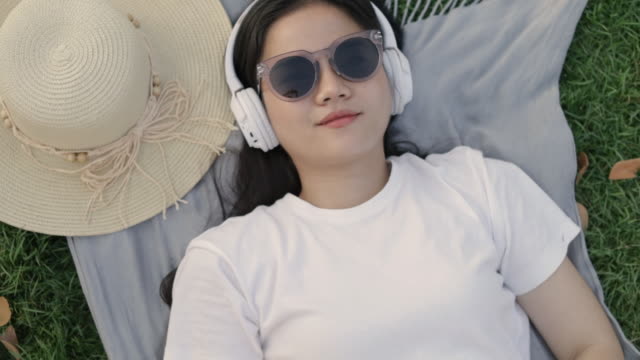 Lächeln-Sie-porträtal-asiatische-Frau-trägt-Sonnenbrille-und-Kopfhörer,-während-auf-dem-Boden-Gras-im-Freien-in-einem-öffentlichen-Park-auf-dem-schönen-Sonnenuntergang-liegen.