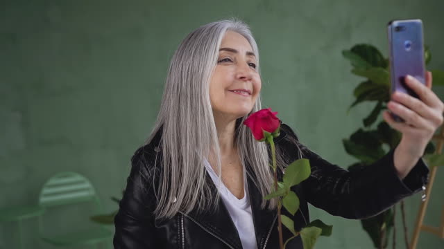 Smiling-satisfied-mature-woman-with-gray-hair-dressed-in-black-jacket-holding-red-rose-in-hand-and-making-selfie