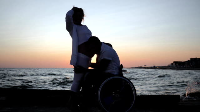 disabled-listens-abdomen-of-his-wife-in-evening,-invalid-spouse-in-wheelchair-with-pregnant-female-at-sunset