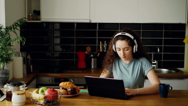 positive-happy-caucasian-pretty-curly-teenage-girl-sitting-at-table-chatting-with-someone-on-laptop-listening-to-music-in-bug-headphones-in-kitchen-indoors