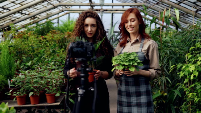 Fröhliche-weibliche-Gärtnerinnen-in-Schürzen-sprechen-und-halten-Blumen-während-der-Aufnahme-video-für-Online-Blog-über-Grünpflanzen-mit-Kamera-auf-Stativ.