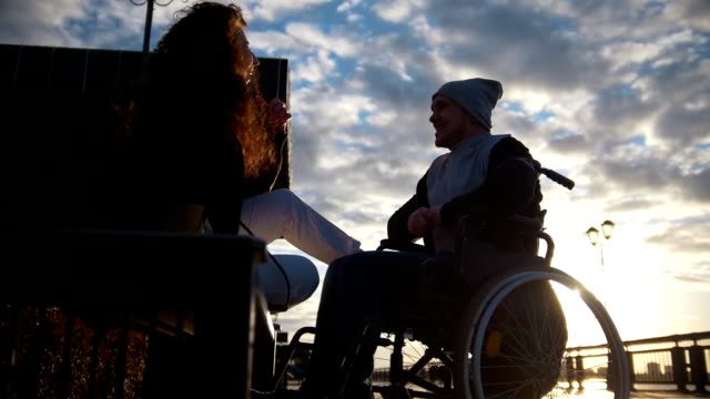 Young-woman-with-disabled-man-in-a-wheelchair-talking-outdoors-at-sunset