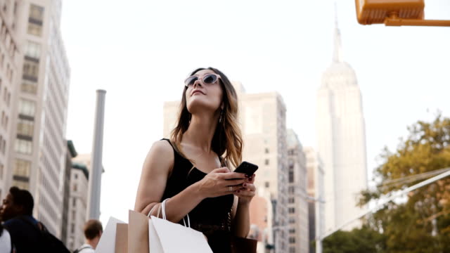 Happy-attractive-Caucasian-young-woman-with-shopping-bags-in-sunglasses-looking-up,-using-smartphone-app-in-New-York
