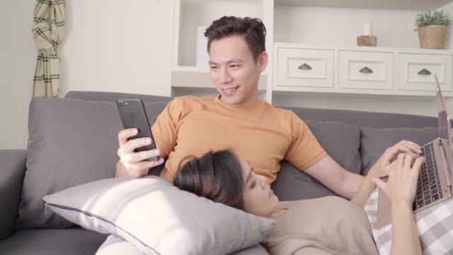 Asian-paar-mit-Smartphone-und-Laptop-Check-social-Media-im-Wohnzimmer-zu-Hause,-süßes-Paar-Liebe-Moment-beim-liegen-auf-dem-Sofa,-wenn-zu-Hause-entspannt-genießen.-Lifestyle-paar-entspannen-zu-Hause-Konzept.