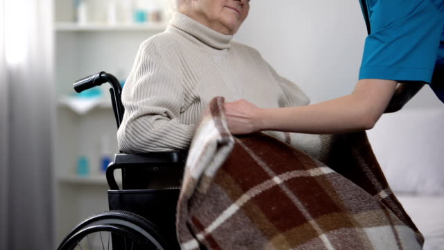 Nurse-covering-old-woman-in-wheelchair-with-blanket,-taking-care-about-patient