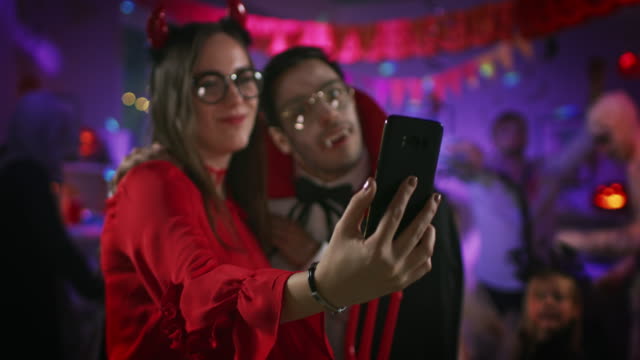 Halloween-Kostümparty:-Verführerische-She-Devil-und-Handsome-Count-Dracula-nehmen-Selfie-für-soziale-Netzwerke-mit-einem-Smartphone.-In-the-Background-Group-of-Monsters-Having-Fun,-Dancing-Under-Disco-Ball.
