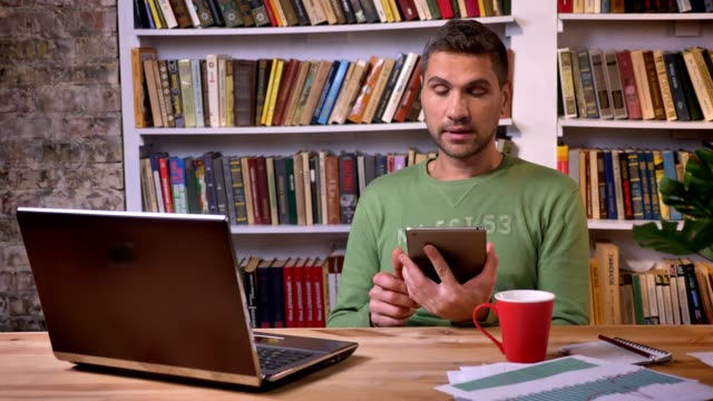 Businessman-sitting-in-front-of-the-laptop-and-having-conversation-on-a-tablet.-Bookshelves-on-the-background