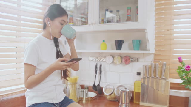 Young-Asian-teenager-woman-drinking-warm-coffee-using-smartphone-listening-music-and-checking-social-in-kitchen-at-home.-Lifestyle-woman-relax-in-morning-at-home-concept.