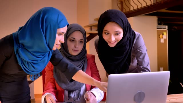 Closeup-portrait-of-three-young-cheerful-muslim-businesswomen-in-hijabs-having-a-lively-conversation-comparing-a-subject-on-the-laptop-on-the-tablet