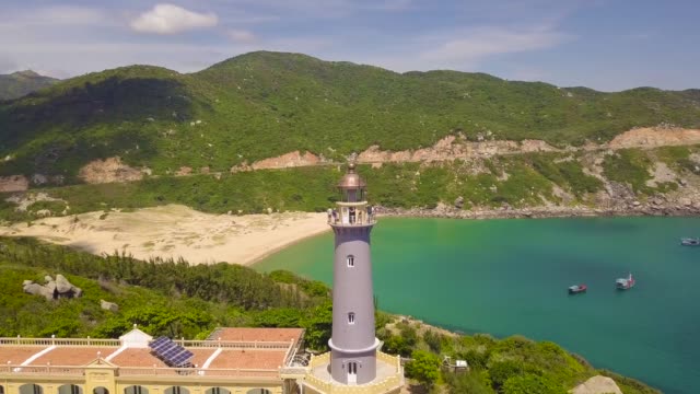 Lighthouse-on-green-mountains-and-ships-sailing-in-blue-sea-aerial-view.-Landscape-from-above-flying-drone-lighthouse-on-tall-mountain-cliff-and-boat-sailing-in-turquoise-sea