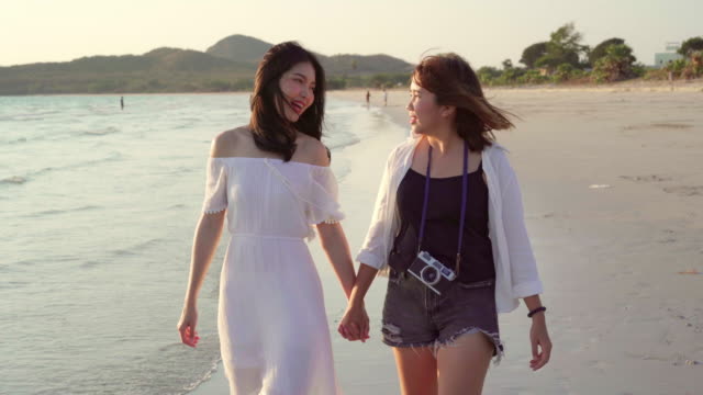 Junges-asiatisches-lesbisches-Paar-zu-Fuß-am-Strand.-Schöne-Freundinnen-freuen-sich-beim-Sonnenuntergang-am-Abend-beim-Spaziergang-am-Strand-am-Meer.-Lifestyle-lesbisches-Ehepaar-reist-nach-Strandkonzept