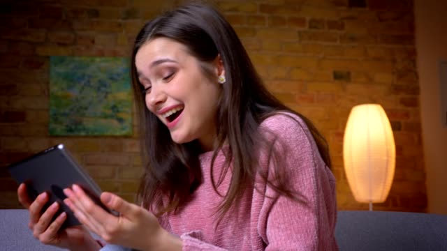 Closeup-shoot-of-young-pretty-brunette-caucasian-female-browsing-on-the-tablet-and-smiling-happily-in-a-cozy-apartment-indoors