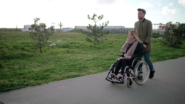 Man-is-rolling-wheelchair-with-young-blonde-woman,-walking-together-in-park