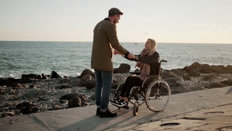 Joyful-man-is-holding-hands-of-his-disabled-girlfriend-on-embankment-of-sea