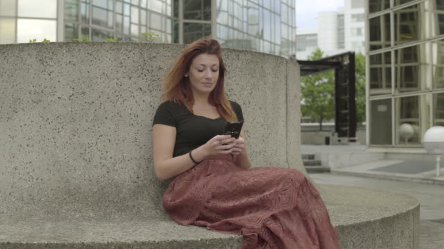 Attractive-woman-using-smartphone-Sitting-in-business-district,-laughing-while-texting-with-mobile-telephone-outside,-typing-text-message-on-cell-phone.-Paris,-Red-hair.-Social-media.-Smiling.-4K.