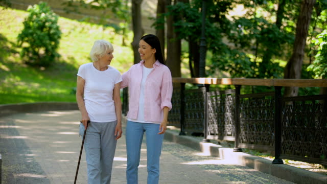 Granddaughter-talking-to-aged-woman-walking-with-stick-in-park,-old-age-health