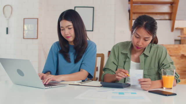 Pareja-lesbiana-asiática-de-negocios-usando-presupuesto-récord-calculadora,-impuestos,-documento-financiero-en-computadora-portátil-trabajando-en-la-cocina.