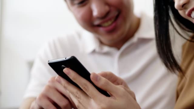 Cierre-la-pareja-asiática-usando-la-comunicación-de-medios-sociales-de-navegación-por-teléfonos-inteligentes-y-las-compras-en-línea-mientras-se-sienta-en-el-sofá-en-casa.